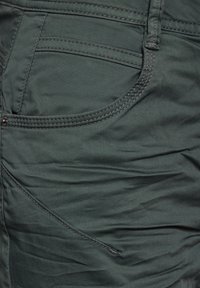 Tecido verde oscuro con una textura suave. Presenta un bolsillo frontal con acentos de costura y una apariencia ligeramente arrugada.