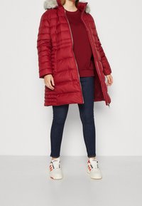 Persoon in een lange rode pufferjas met een bontkap, bordeaux trui, donkere skinny jeans en witte Adidas sneakers met rode strepen.