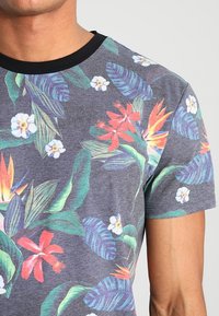 Camiseta de estampado floral tropical con hojas verdes y moradas, flores rojas y naranjas, flores blancas y un escote redondo negro.