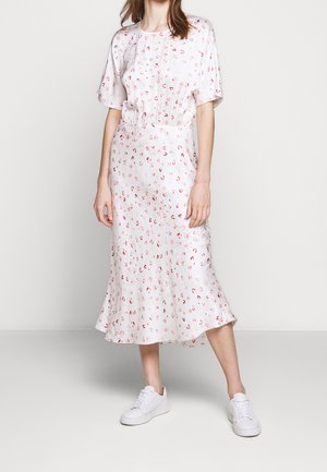 Femme portant une robe midi blanche avec de petits imprimés abstraits roses et rouges, associée à des baskets blanches, debout devant un fond uni.