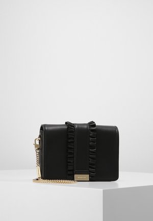 Sac bandoulière en cuir noir avec sangle avant à volants, chaîne dorée et plaque de marque dorée, présenté sur une surface blanche.