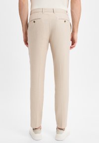 Pantaloni in lino beige con una superficie leggermente testurizzata, vestibilità affusolata e due tasche posteriori con chiusura a bottone. Indossati con sneakers bianche.