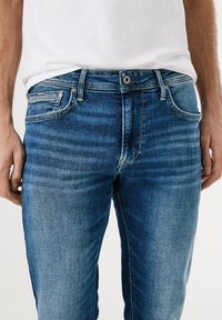 Modri jeans iz denima s klasičnim dizajnom, vidnimi šivi, petimi žepi, kovinsko nitenje in gumbom za zapiranje.