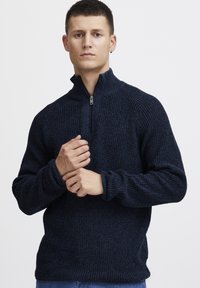 Pulover tricotat de culoare navy, cu guler înalt și fermoar pe jumătate. Are un model texturat și mâneci lungi cu manșete ribbate.