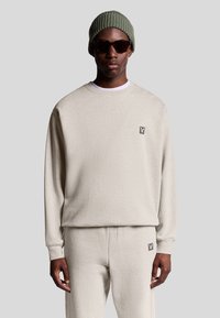 Uomo in felpa beige e pantaloni coordinati con piccole toppe logate, berretto in maglia verde e occhiali da sole scuri su uno sfondo neutro.
