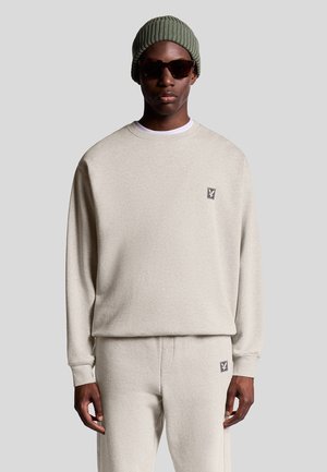Mand iført beige sweatshirt og matchende bukser med små logo-patch, grøn strikket beanie og mørke solbriller mod en ensfarvet baggrund.