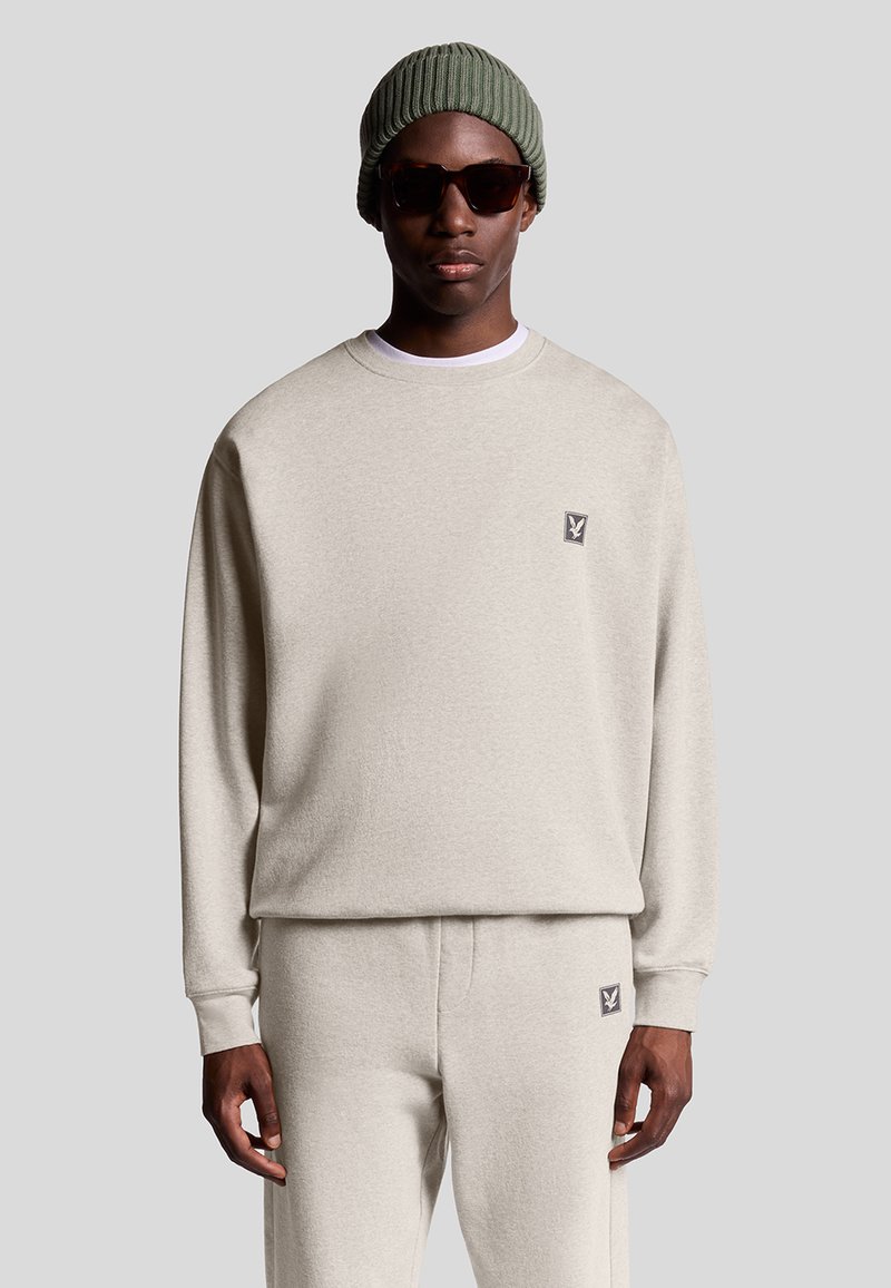Uomo in felpa beige e pantaloni coordinati con piccole toppe logate, berretto in maglia verde e occhiali da sole scuri su uno sfondo neutro.