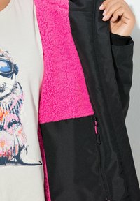 Chaqueta negra con un interior de forro polar rosa brillante, que cuenta con un bolsillo con cremallera y una tela texturizada. Diseño en capas con colores contrastantes.