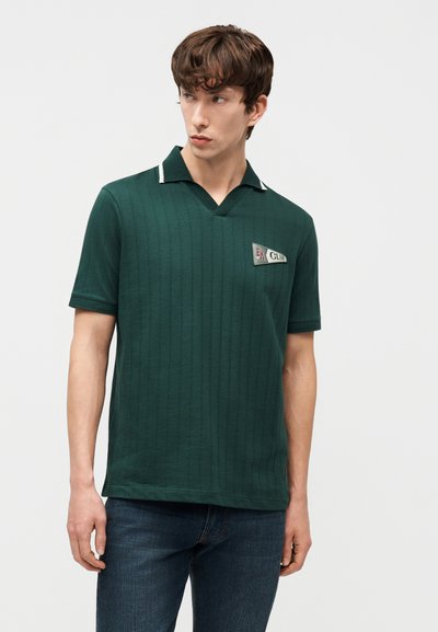 Νεαρός άνδρας που φοράει σκούρο πράσινο πόλο μπλουζάκι με κοντά μανίκια και ένα μικρό έμβλημα «EA Club» και σκούρο μπλε τζιν, κοιτάζοντας προς τα αριστερά.