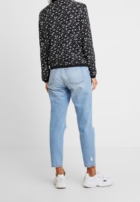 Veste bomber noire avec motif floral blanc, associée à un jean bleu clair déchiré et des baskets blanches. Vue de dos ; poignets et ourlet élastiques.