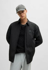 Black button-up jacket with a textured knit sweater beneath, complemented by a gray cap and light gray trousers. Features logo detail.