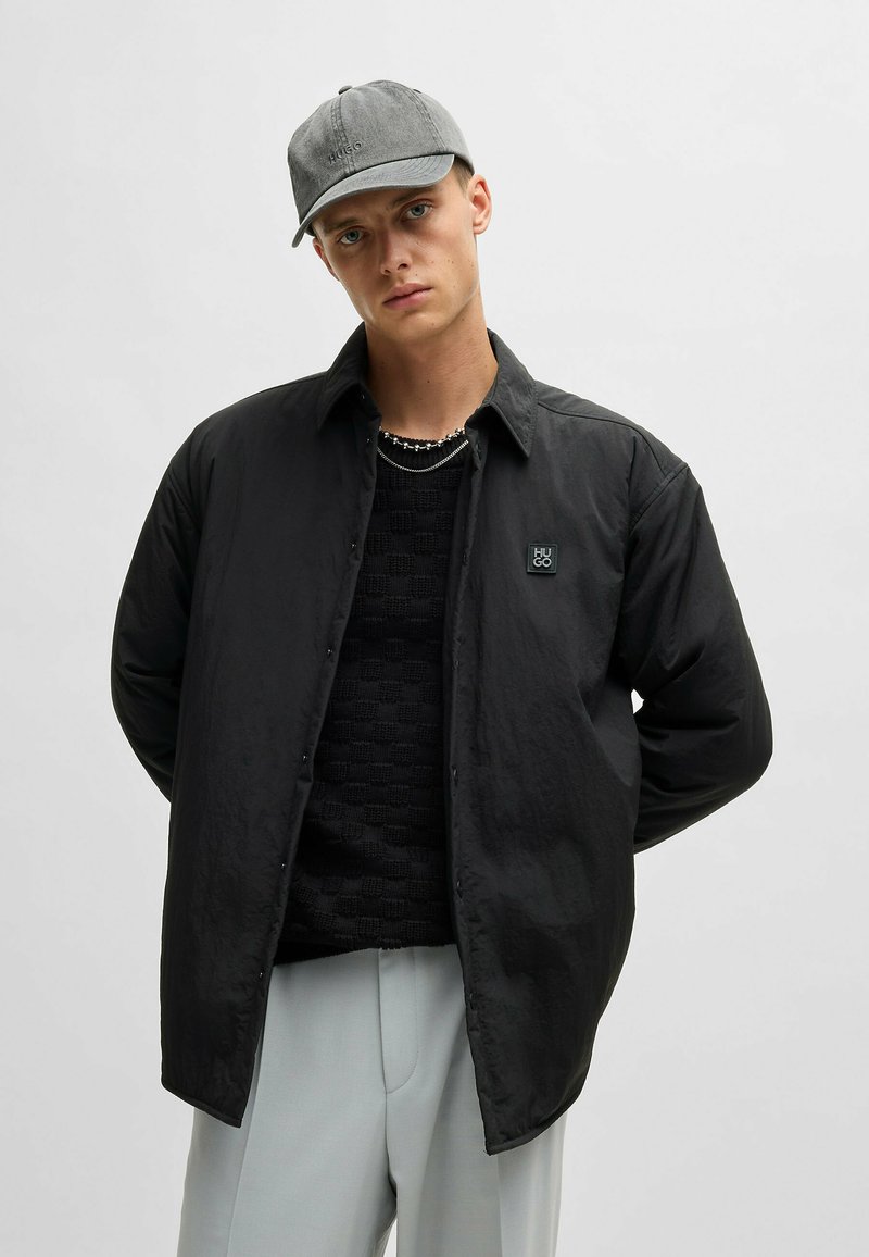 Black button-up jacket with a textured knit sweater beneath, complemented by a gray cap and light gray trousers. Features logo detail.