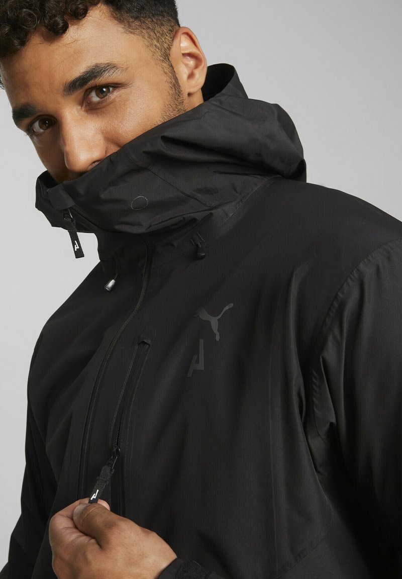 Schwarze isolierte Jacke mit hohem Kragen, Reißverschluss, Logopunkten und glatter Textur. Verfügt über verstellbare Kordeln und Taschen.