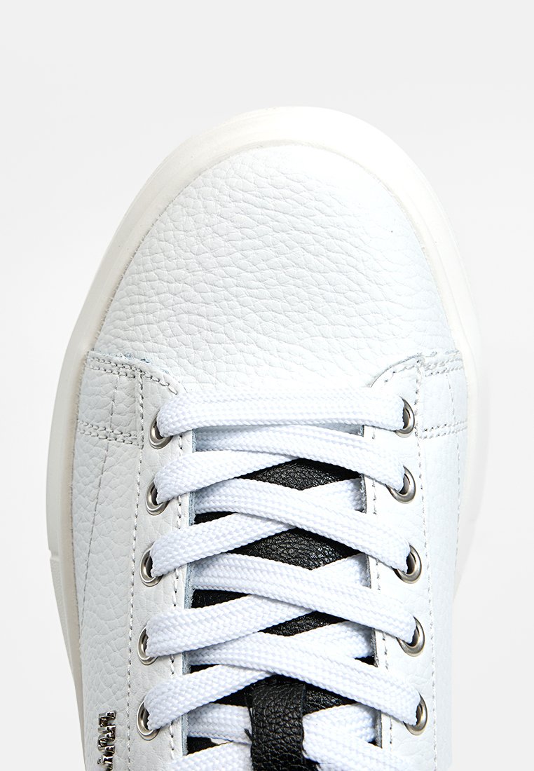 Zapatilla de cuero blanca con acabado texturizado, panel de encaje negro y cordones blancos. Presenta ojales plateados y un diseño de puntera redondeada.