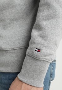 Primo piano di una persona che indossa una felpa grigia con un piccolo logo rettangolare rosso, bianco e blu navy sul polsino della manica.