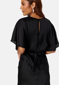 Zwarte satijnen jurk met korte, doorschijnende mouwen; de achterkant heeft een kleine knoopsluiting en een keyhole-opening onder de halslijn.