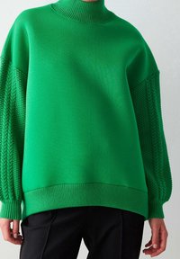 Suéter verde de gran tamaño con cuello alto, que presenta mangas de canalé texturizadas y un cuerpo sólido. Fabricado en un material suave y elástico.