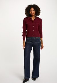 Burgundy stickad kofta med guldknappar, två bröstfickor, ihopparad med mörka denim jeans med vida ben och kontrasterande sömmar.