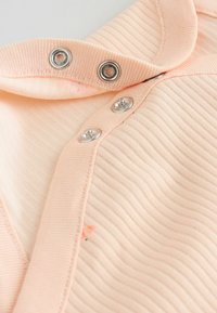 Gros plan sur un tissu côtelé rose clair avec trois boutons-pression métalliques partiellement attachés sur une ouverture de vêtement.