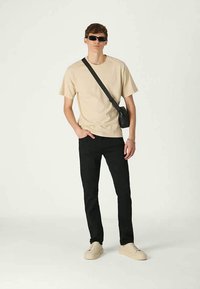 Young man wearing black sunglasses, beige t-shirt, black pants, beige sneakers, and a black crossbody bag, standing with one hand in pocket.