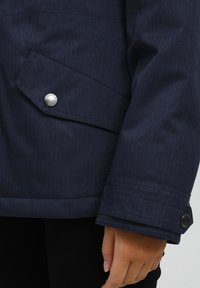Marineblauwe herringbone-gepatroonde jas met een zijzak, zilveren drukknoop en gedetailleerde stiksels bij de manchetten.