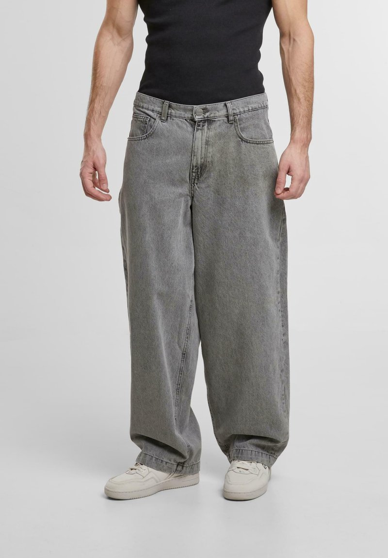Jean baggy gris clair avec une texture usée, présentant de grandes poches latérales et une fermeture à bouton à la taille, assortis à des baskets blanches.