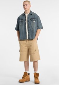 Denim button-up shirt met borstzakken, tan cargo shorts en bruine werklaarzen. Beschikt over een relaxte pasvorm en korte mouwen.