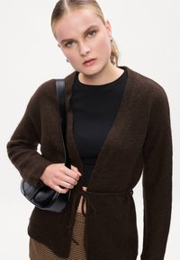 Cardigan marrón de punto con diseño envolvente, mangas largas y cierre con lazo. Llevado sobre una blusa negra ajustada, combinado con un bolso pequeño negro.