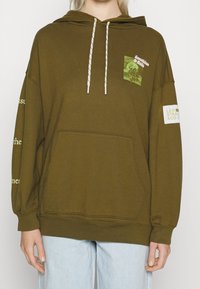 Hoodie vert olive avec une poche avant, cordons blancs et motif sur le côté gauche de la poitrine. Texte sur les manches et écusson logo sur la droite.