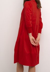 Abito rosso con maniche a sbuffo e una vestibilità comoda, caratterizzato da una superficie liscia e da un leggero lucore. Lunghezza al ginocchio con dettaglio in vita arricciato.