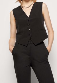 Schwarze ärmellose Weste mit V-Ausschnitt, fünf Knöpfen vorne und zwei seitlichen Taschen; aus glattem Material mit tailliertem Schnitt.