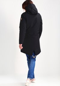 Svart lång jacka med huva, med en delad bakdesign, texturerat tyg och detaljer i guldmetall. Bärs över blå jeans.