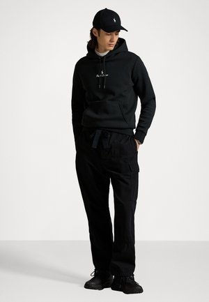 Felpa nera con cappuccio, cordino e logo, abbinata a pantaloni cargo neri con tasche e sneakers nere. Abbigliamento sportivo semplice e casual.