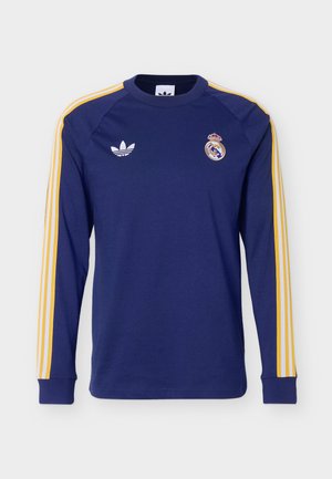 Maglia a maniche lunghe blu navy con strisce gialle sulle maniche, logo Adidas sul petto sinistro ed emblema del Real Madrid sul petto destro.
