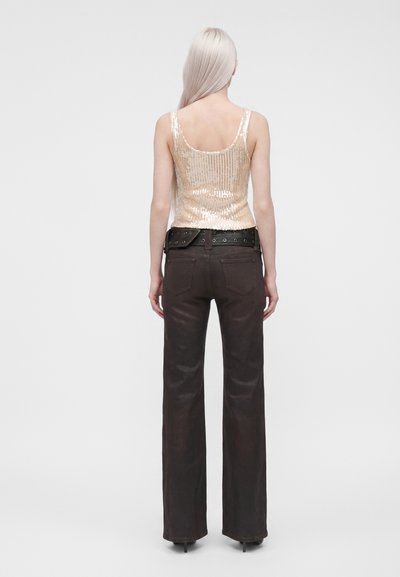 Femme aux longs cheveux platine, de dos, portant un haut sans manches doré à sequins, une ceinture noire et un pantalon ample foncé avec des talons.