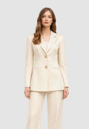Donna che indossa un blazer sartoriale color crema con due bottoni e pantaloni a gamba larga abbinati, su uno sfondo chiaro e semplice.