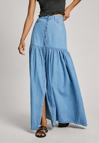 Ljusblå denimmaxikjol med lagerdesign, med knappar fram och en sidoklyvning, kombinerad med svarta sandaler.