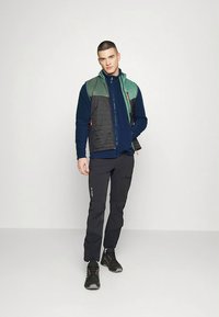Men's outdoor outfit featuring a padded vest in green and black, a navy fleece underneath, and black pants. Includes dark footwear.
