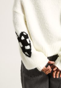 Maglione bianco lavorato a maglia con design a macchie nere sulla manica sinistra, polsini a costine e texture morbida. Abbinato a pantaloni grigio scuro.