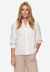 Witte blouse met knopen, kleine blauwe bloemenprint, geplooide textuur en pofmouwen. Gecombineerd met lichtbeige wijde broekspijpen.