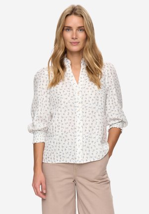 Weiße Hemdbluse mit kleinem blauem Blumenmuster, plissierter Textur und Puffärmeln. Kombiniert mit hellbeigen, weit geschnittenen Hosen.