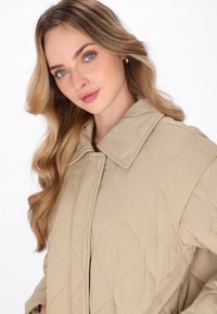 Beige quiltet frakke med stor krave og teksturerede bølge mønstre. Har en frontlynlås og blødt stof. Ingen åbenlyse hardwaredetaljer.