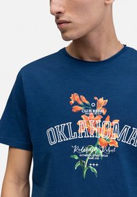 Camiseta de manga corta azul marino con un gráfico floral naranja que incluye el texto "OKLAHOMA" y "CLUB REBEL", con cuello redondo y textura suave.