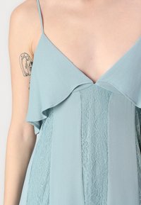 Svetlo modra obleka z V-izrezom, večplastnimi volančki na rokavih in čipkastimi detajli, z gladko, tekočo teksturo in prilegajočim pasom.