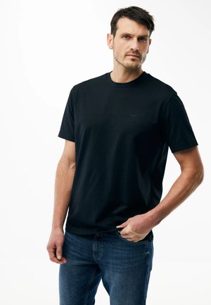 Hombre vestido con camiseta negra de manga corta y pantalones vaqueros azules, de pie con una mano en el bolsillo contra un fondo blanco.