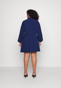 Sininen ruudullinen mekko, jossa on muodokas vyötärö ja puffahtavat hihat, suora helma ja shortsit. Pukeutuu mustiin korkokenkiin.
