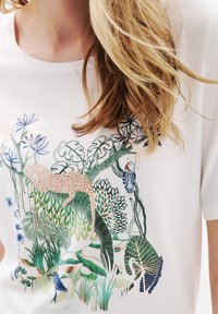 Camiseta blanca con bordado colorido de temática selvática, que incluye un leopardo y hojas tropicales en tonos verdes, azules y coral.