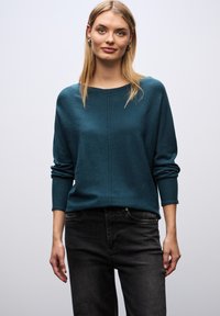 Turkusowy sweter z okrągłym dekoltem, szerokimi rękawami i prążkowanymi mankietami, zestawiony z ciemnymi dżinsami. Gładka faktura, minimalistyczny design, casualowy wygląd.