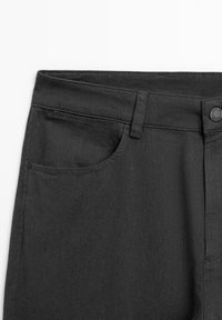 I pantaloni in denim neri presentano una vita standard, una tasca frontale a sinistra e una chiusura con bottone in metallo con una leggera texture visibile.
