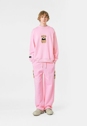 Sudadera rosa con un diseño de oso, combinada con pantalones rosas que presentan bolsillos laterales. La tela parece suave y casual, con un corte relajado.